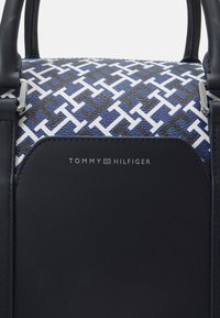 Kabelka Tommy Hilfiger s texturovaným tmavě modrým základem, bílým vzorem loga, černými koženými detaily a stříbrnými kovovými prvky.