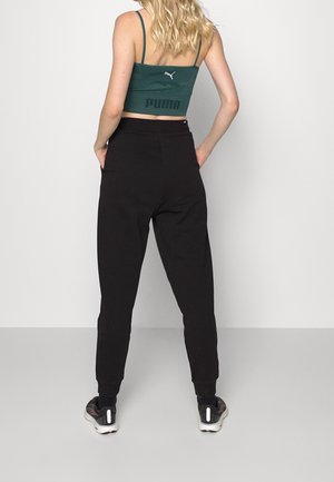 Frau mit blonden Haaren steht mit den Händen in den Taschen, trägt ein grünes Puma-Crop-Top, schwarze Hose und schwarze Sportschuhe vor einem einfarbigen Hintergrund.