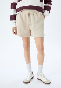 Beige shorts puuvillast, millel on elastne vöökoht ja pael, külgedel taskud ning lahti istuv lõige, kantud valgete tossude ja sokkidega.