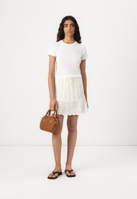 Camiseta blanca de manga corta combinada con una falda de croché color crema, sosteniendo un pequeño bolso marrón texturizado. Sandalias planas negras completan el conjunto.