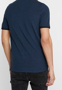 Man som bär en tight, marinblå kortärmad T-shirt och svarta jeans, visad bakifrån mot en ljus enkel bakgrund.