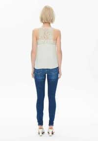 Top leggero senza maniche con schiena in pizzo, abbinato a jeans blu aderenti. Modella che indossa tacchi alti beige, in piedi davanti a uno sfondo semplice.