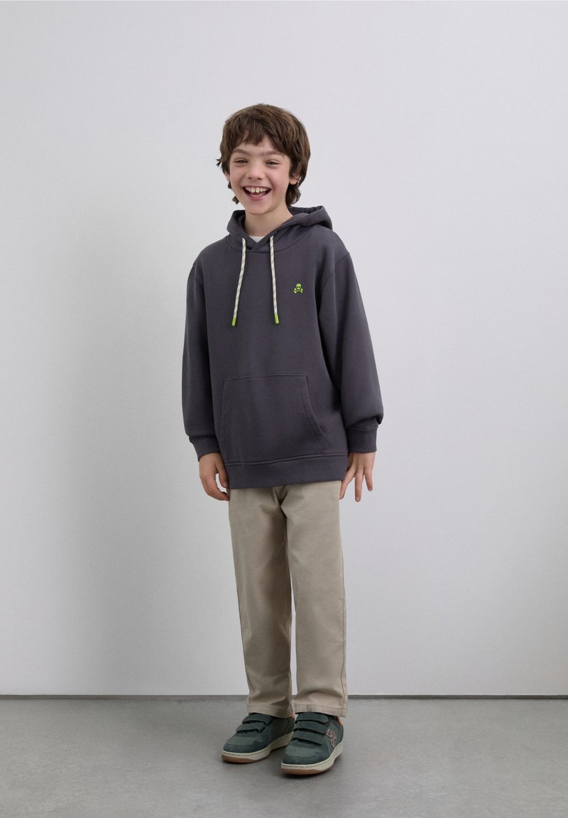 Gray hoodie with green accent logo, hood drawstrings, front pocket, paired with beige pants and teal shoes. Simple, casual outfit.