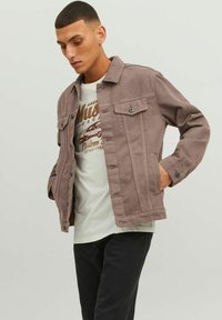 Giacca in cotone grigia con chiusura a bottoni, tasche sul petto e vestibilità rilassata, indossata sopra una t-shirt chiara, abbinata a jeans scuri.