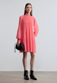 Abito rosa plissettato con maniche lunghe, collo alto, design fluente, abbinato a una borsa nera e stivaletti alla caviglia. Tessuto liscio.