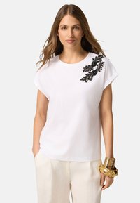 Camiseta blanca de manga corta con un adorno floral de cuentas y lentejuelas negras en el hombro izquierdo, tejido de algodón suave, corte relajado.