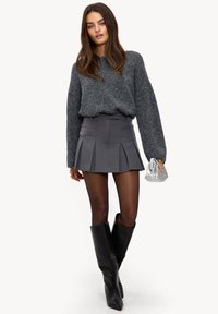 Pull gris duveteux, mini-jupe grise plissée, bottes noires montantes jusqu'aux genoux, tenant un petit sac à main argenté. Textures lisses et coupe ajustée.