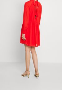 Punainen, reiden pituinen mekko, jossa on pitkät hihat, takana solmittava yksityiskohta ja rypytetty vyötärö. Asusteena nudet, nyörilliset korkokengät.