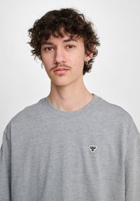 Sudadera gris con cuello redondo, de textura suave y mangas cortas. Presenta un pequeño logo negro en el lado izquierdo del pecho.