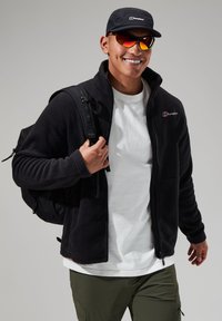 Black fleece jacket with zip, worn over a white t-shirt. Matching black backpack, and black cap. Red and yellow sunglasses complete the outfit.