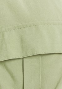 Tessuto verde chiaro con una texture liscia, caratterizzato da un orlo piegato e pieghe sottili per un dettaglio di design. La cucitura è eseguita con precisione.