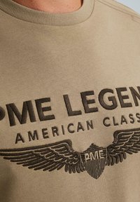 Bežová tkanina s čierne vyšívaným textom "PME Legend American Classic" a logom s krídlovou štítou obsahujúcim písmená "PME".