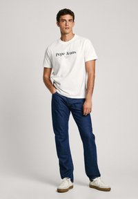Camiseta de algodón blanca con el logo negro de "Pepe Jeans", combinada con jeans de mezclilla azules y zapatillas blancas, con un corte clásico y cuello redondo.