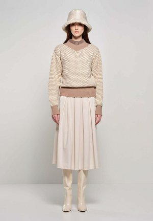 Vrouw draagt een gestructureerde beige trui, een crèmekleurige geplooide rok, crèmekleurige kniehoge laarzen en een donzige beige bucket hat tegen een effen achtergrond.