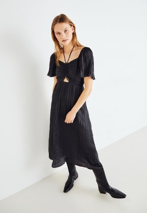 Robe noire à mi-mollet avec un buste froncé, des manches courtes bouffantes et un détail découpé à la taille. Texture de tissu lisse et froissé.