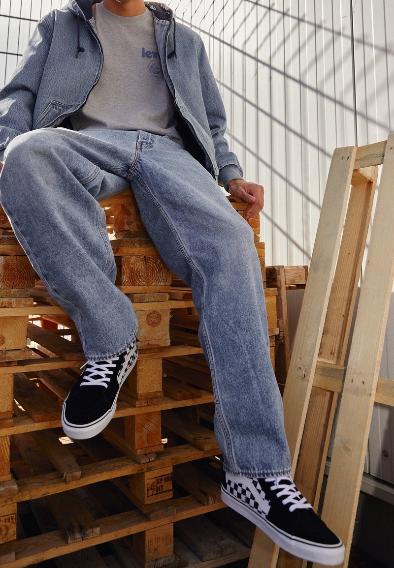 Veste en jean et jeans bleu clair, t-shirt gris, baskets à carreaux noir et blanc. Modèle assis sur des palettes en bois.