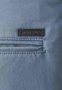 Jasnoniebieska dżinsowa tkanina z wyszywaną kieszenią. Czarna skórzana metka z wytłoczonym napisem "JACK & JONES". Gładka tekstura, jednolity kolor.