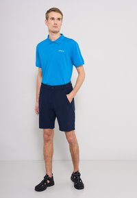 Blå polotröja med krage och tre knappar, matchad med marinblå shorts med elastisk midja och sidofickor. Svarta sandaler med utskärningar.