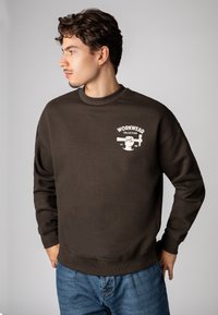 Brown sweatshirt with a crew neck and ribbed cuffs, featuring a white graphic on the chest. Paired with blue jeans.