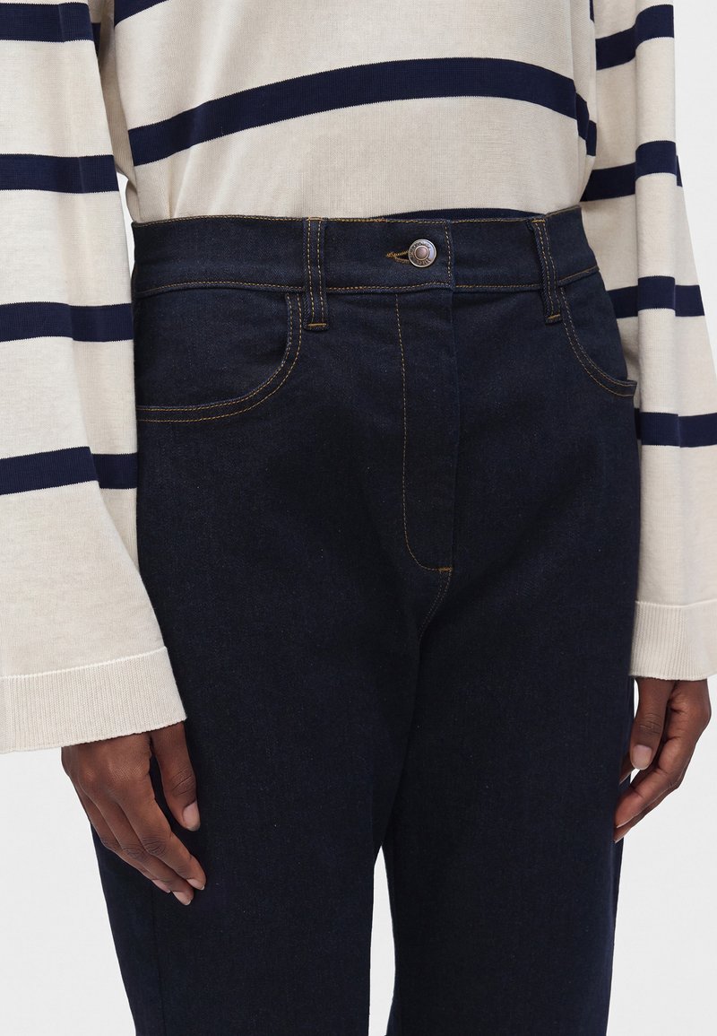 Gros plan d'une personne portant un jean en denim bleu foncé avec des surpiqûres jaunes et une chemise crème à manches longues avec des rayures horizontales bleu marine.