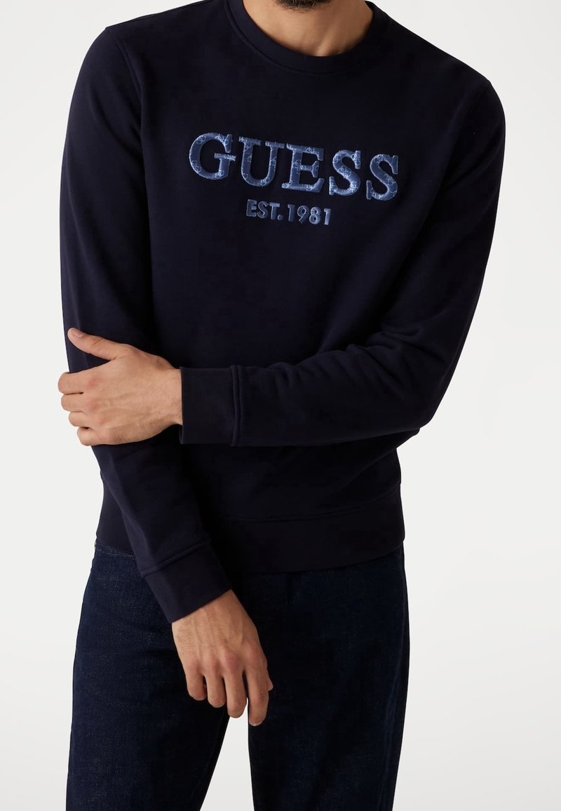 Homme portant un sweat-shirt noir avec l'inscription bleue « GUESS EST. 1981 », associé à un jean bleu foncé sur fond blanc.