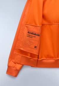 Giacca sportiva arancione con tessuto testurizzato, polsini a costine e una tasca con un'etichetta che dettaglia la "Circular Series" e un codice QR.