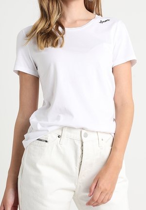 Kvinde iført en ensfarvet hvid T-shirt med teksten "Amore" nær kraven og højtaljede hvide bukser med knapper.