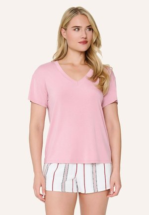 Žena s dlhými blond vlasmi, ktorá nosí svetloružové tričko s výstrihom do V a biele šortky s vertikálnymi červenými a sivými pruhmi, stojaca pred jednoduchým pozadím.