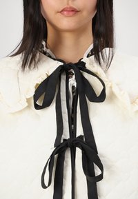 Κουβερτίνα ιβουάρ με ραφές και ριγέ γιακά, μαύρες μεταξωτές κορδέλες στον λαιμό και εσωτερική επένδυση με λουλουδάτο σχέδιο. Ορατή λεπτομέρεια ραφής σε σχήμα καρδιάς.