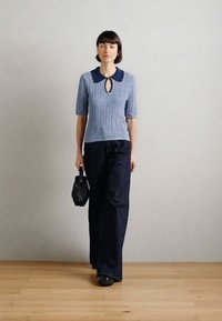 Ανοιχτό μπλε πλεχτό τοπ με σκούρο μπλε γιακά και λεπτομέρεια κλειδαρότρυπας, συνδυασμένο με σκούρα τζην παντελόνια; κρατώντας μια μικρή μαύρη τσάντα.