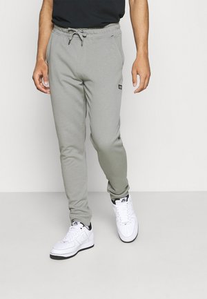 Mężczyzna ubrany w jasnoszare spodnie dresowe ze ściągaczem w pasie i sznurowadłami oraz białe buty Nike, stojący na tle jednolitego białego tła.