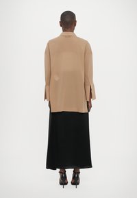 Beige oversized shirt with long sleeves and side slits, paired with a long black skirt, worn with black heeled shoes.