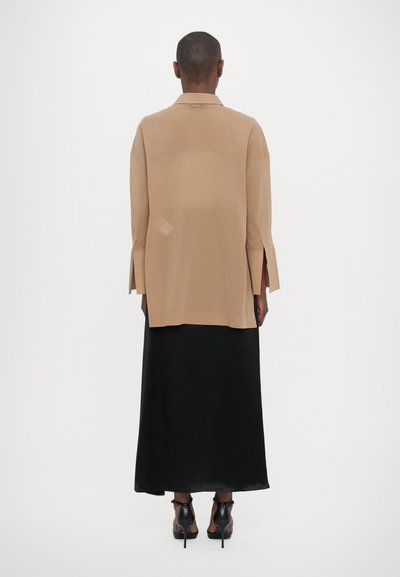 Beige ylimitoitu paita pitkillä hihoilla ja sivuaukkoilla, yhdistettynä pitkään mustaan hameeseen, käytetään mustien korkokenkien kanssa.