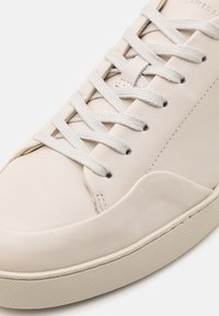 Beżowe skórzane sneakersy z płaskimi białymi sznurówkami, okrągłym noskiem i teksturowaną gumową podeszwą. Charakteryzują się subtelnym przeszyciem i gładką powierzchnią.