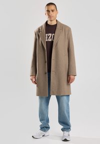 Cappotto di lana marrone con vestibilità su misura, ampi rever e taglio dritto. Indossato sopra un felpa scura e abbinato a jeans azzurri chiari e sneakers.