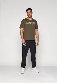 Vyras, dėvintis olivinės spalvos UFC logotipo marškinėlius, juodus kelnes su logotipu ir baltus sportinius batelius, stovi prieš paprastą šviesų foną.