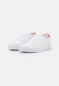 Sneakers bianche con lacci, dalla superficie liscia, punta rotonda e dettagli rossi sul tallone. Presentano un particolare logo in rilievo sul lato e una suola in gomma testurizzata.