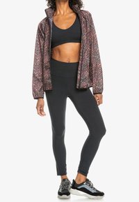 Veste noire courte avec un motif floral rose, brassière de sport noire, leggings noirs taille haute, et chaussures de sport noires avec des accents gris.