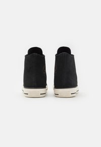 Černé semišové kotníkové boty s texturovaným povrchem, nesoucí bílou gumovou podrážku a černé pruhované akcenty. Vytlačené logo Converse na straně.