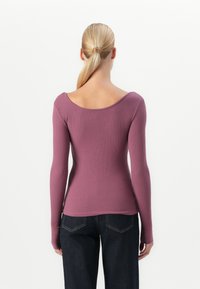 Kvinne med blond hestehale iført en tettsittende, langermet ribbestrikket mauve genser og mørkeblå denimjeans, stående med ryggen til.