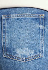 Poche arrière d'un jean en denim bleu avec des coutures orange et un chemise blanche légèrement visible au-dessus de la ceinture.