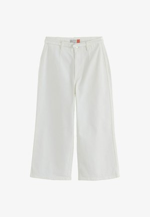 Femeie purtând pantaloni albi cu talie înaltă, cu o curea subțire și un top scurt din denim albastru, mâna odihnindu-se lângă talie.