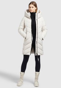 Ljusbeige puffjacka med huva, med en kviltad design, zippered front och sidofickor. Bärs över en svart polotröja och leggings.