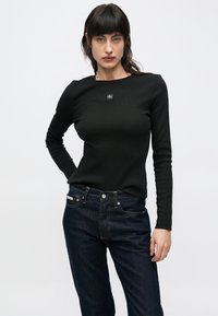 Svart, ribbestrikket genser med lange ermer, rund hals og en liten logopatch midt på; kombinert med mørkeblå denimjeans.