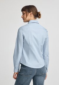 Camicia a maniche lunghe azzurro chiaro con colletto classico, leggera lucentezza, vestibilità aderente e polsini con bottoni, abbinata a jeans scuri.