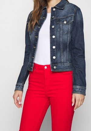 Femme portant une veste en denim bleu foncé sur une chemise blanche, assortie à un pantalon rouge vif, debout devant un fond uni.