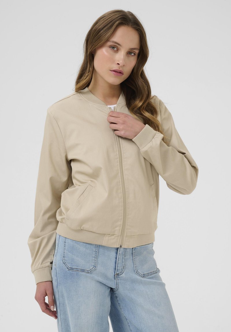 Veste bomber beige clair en tissu lisse, avec une fermeture éclair à l'avant, des poignets côtelés et des poches latérales. Portée par-dessus un jean en denim bleu.