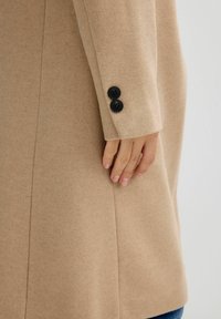 Abrigo de lana beige con un diseño estructurado, de textura suave y dos botones negros en la manga para abrochar.