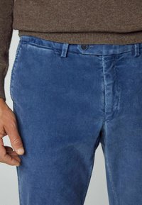 Pantalon en velours côtelé bleu avec taille boutonnée porté avec un pull en maille marron, montrant la main gauche d'une personne reposant près de la cuisse.