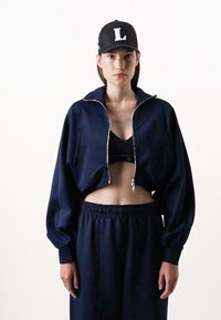 Giacca cropped navy con zip, maniche larghe e due tasche anteriori, abbinata a una maglietta cropped nera e pantaloni larghi coordinati.